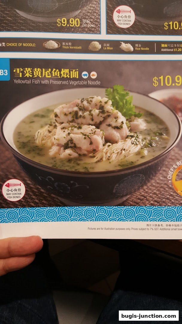 A close-up of a menu item showing a bowl of noodle soup with fish and preserved vegetables.