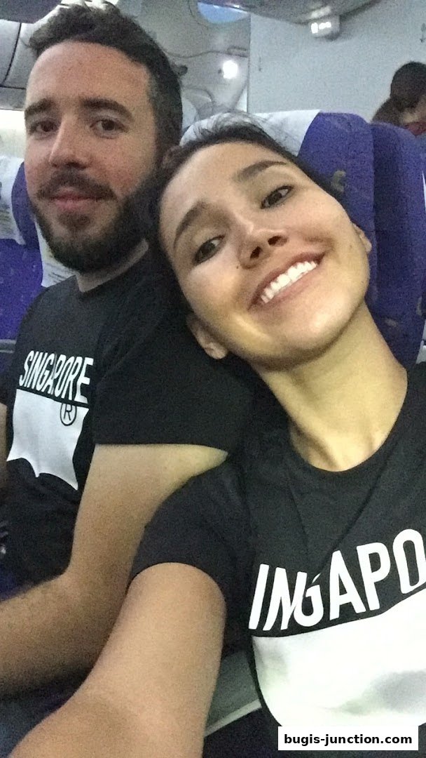 A couple smiles for a selfie on an airplane, both wearing black t-shirts with 'SINGAPORE' printed on them.