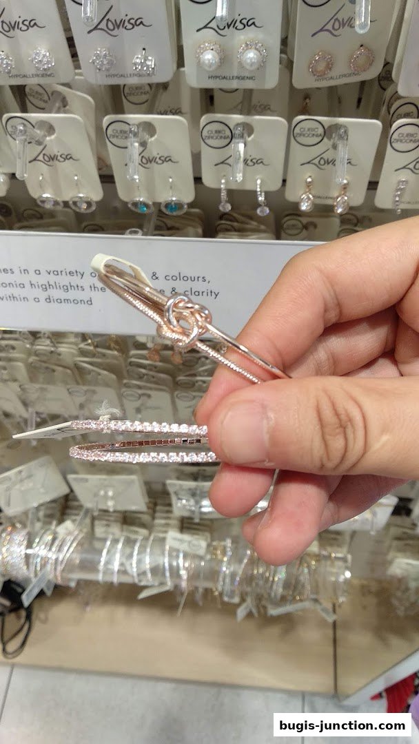 A hand holds rose gold knot bracelet and diamond bracelet in front of a display of earrings.