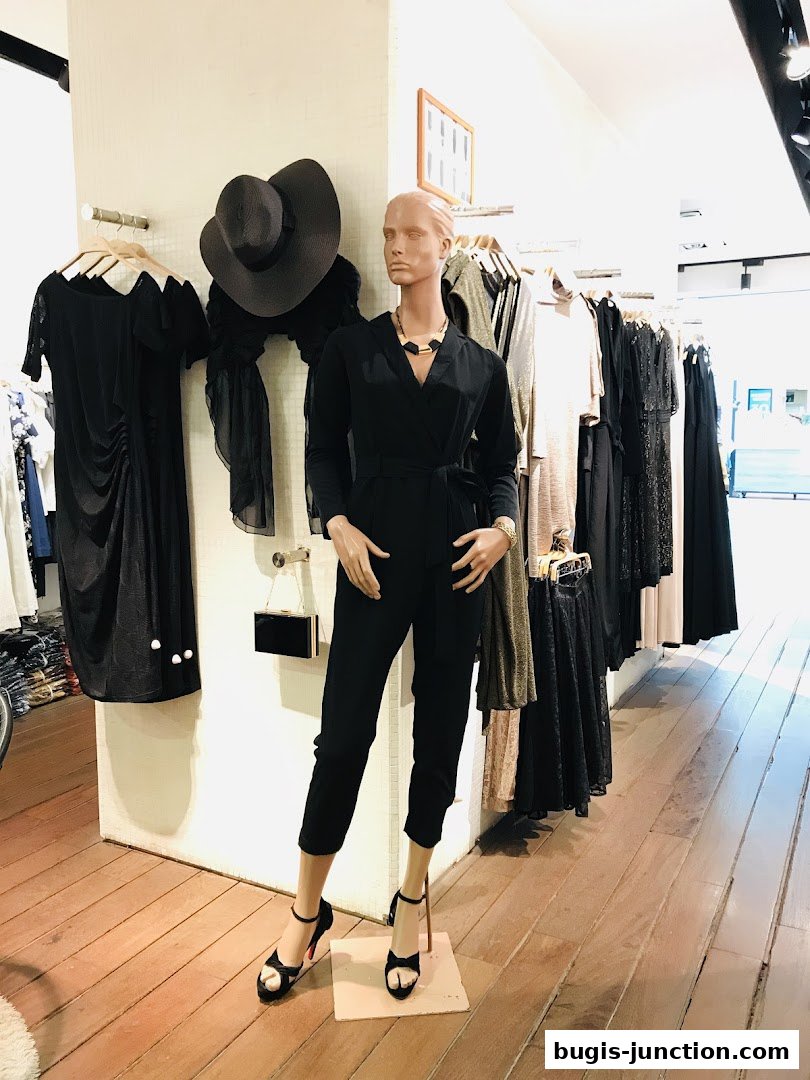A mannequin models a black jumpsuit with a hat and dresses displayed in a boutique.