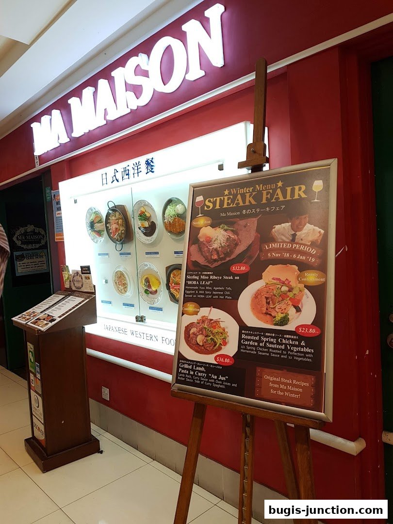 The storefront of Ma Maison restaurant, featuring a prominent sign and a display of food items, with a winter steak fair menu.