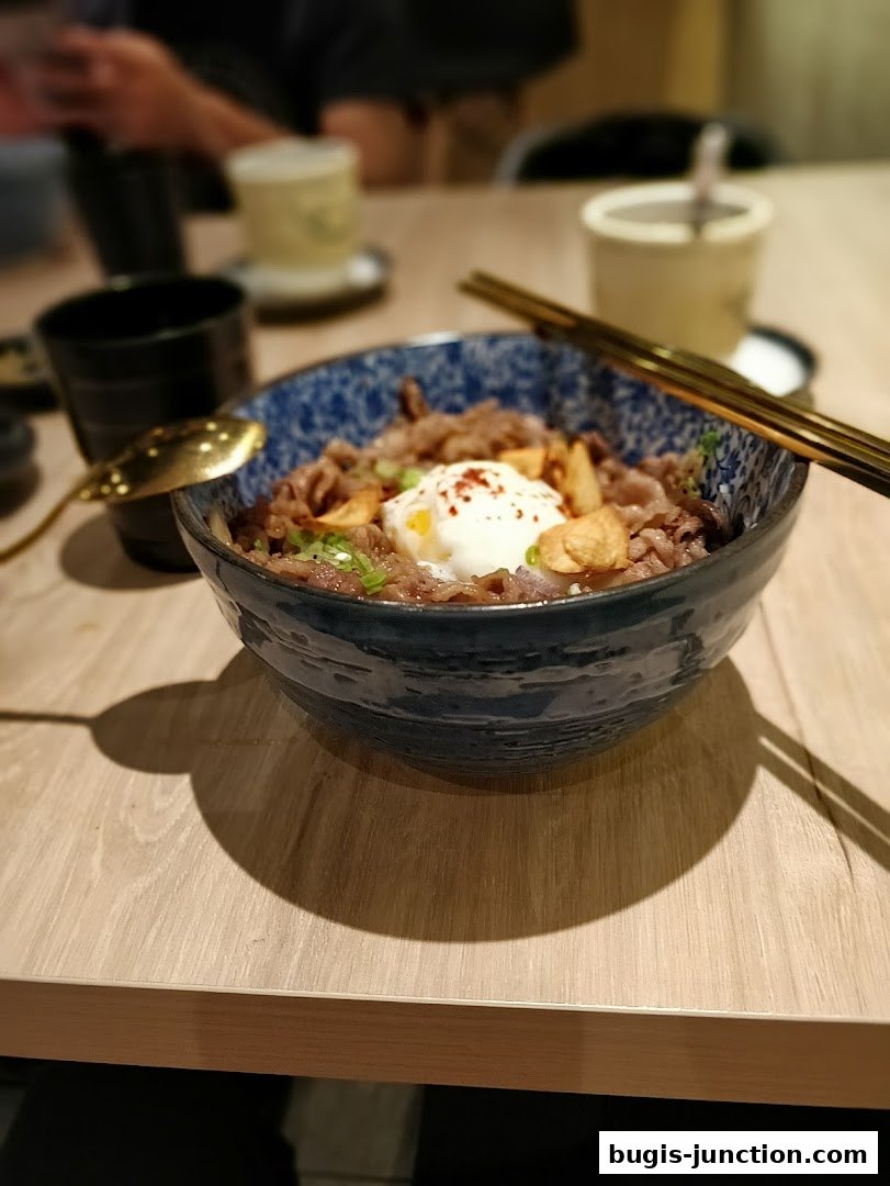 A close-up of a Japanese rice bowl with beef, egg, and garlic chips, served with chopsticks.