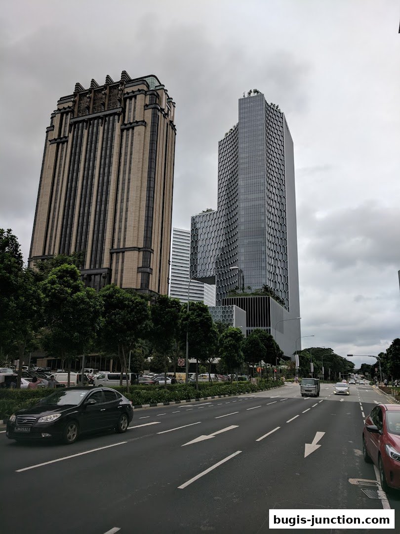 A street view with modern skyscrapers and trees under a cloudy sky.