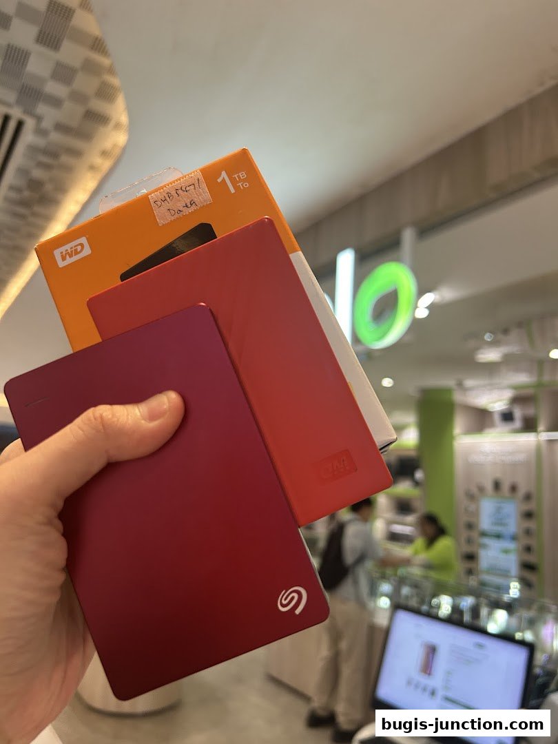 A hand holds several portable hard drives in front of a store with a green logo.