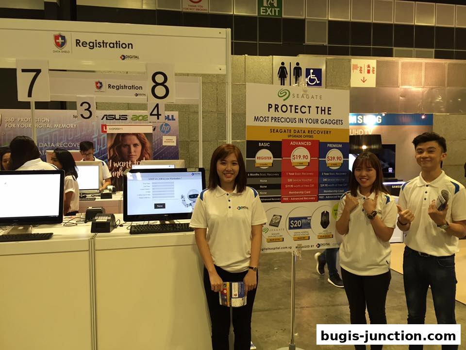 Three smiling staff members stand at a digital hospital registration booth with computers and promotional materials.