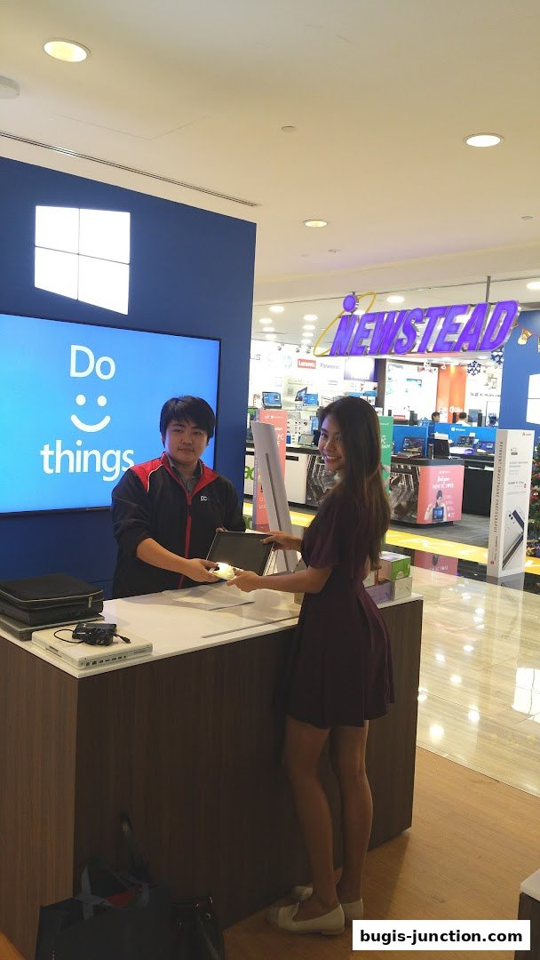 A store employee hands a laptop to a customer at a counter with a Windows logo.