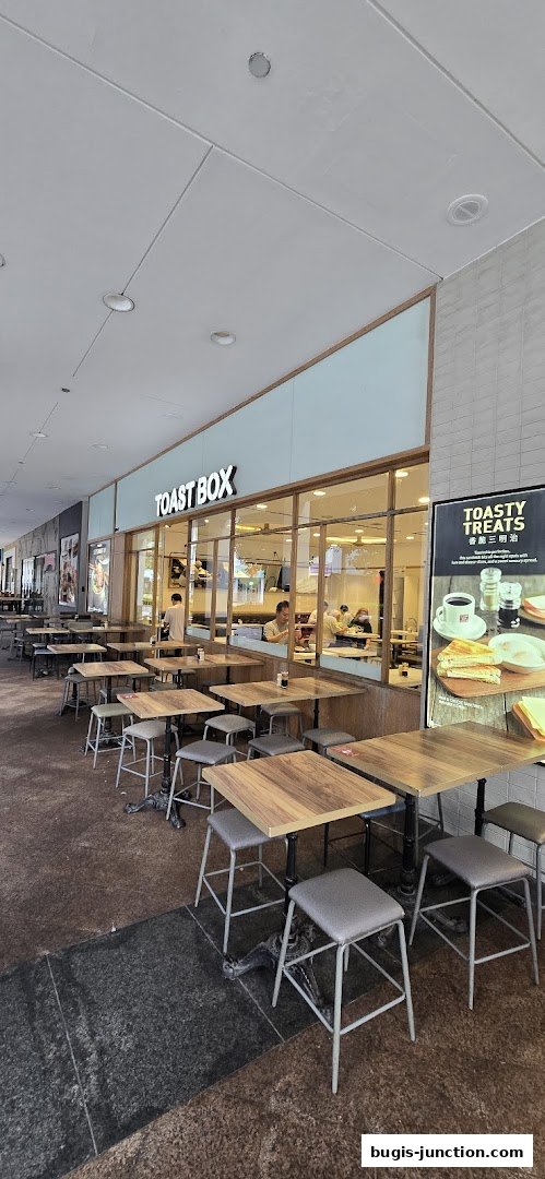 The exterior of a Toast Box cafe with outdoor seating and a promotional poster.