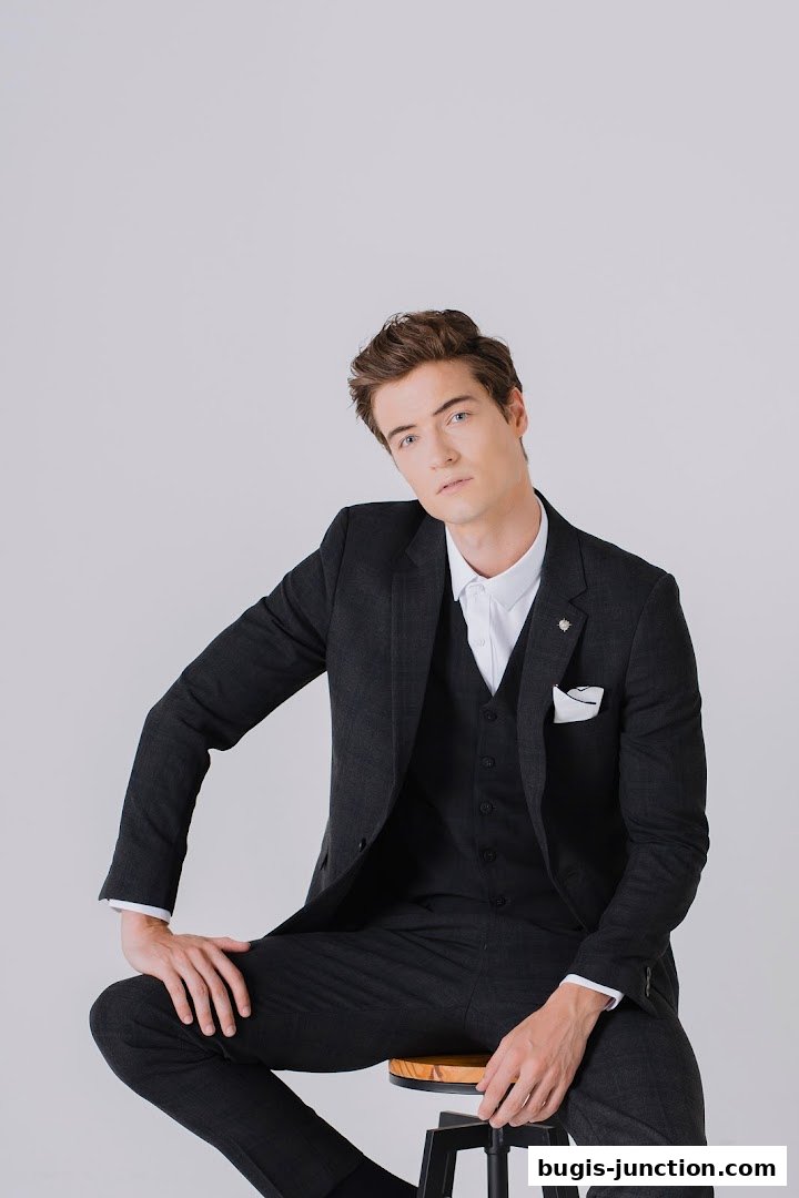 A man in a dark grey suit and white shirt poses on a stool against a white background.