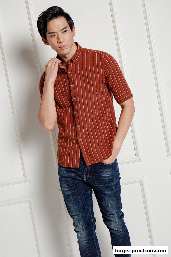 A young man models a striped rust-colored shirt and distressed denim jeans.