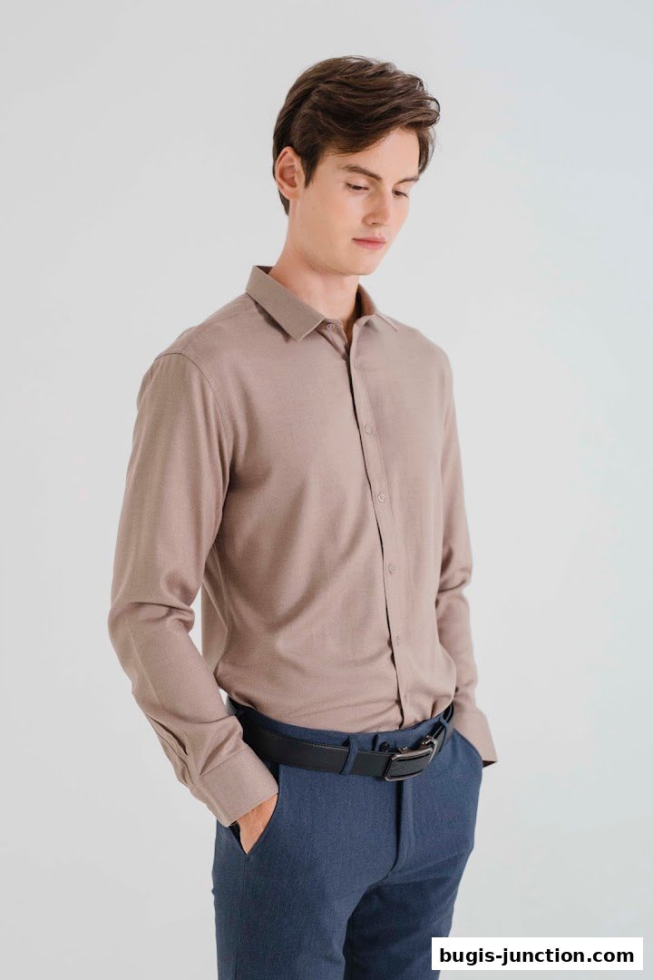 A young man models a light brown collared shirt and blue trousers with a black belt.