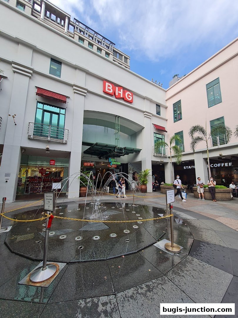 Exterior view of a shopping mall entrance with a fountain and various shops.