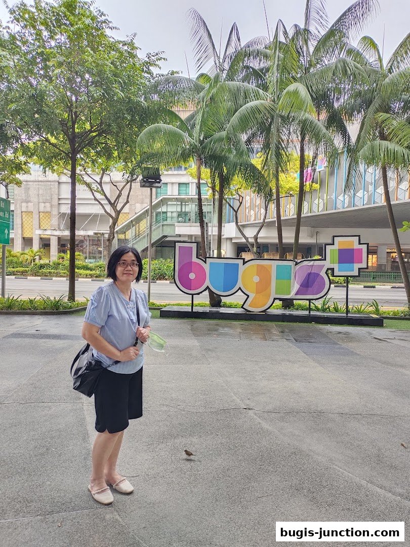 A woman stands in front of a colorful 'Bugis' sign with palm trees and a building in the background.