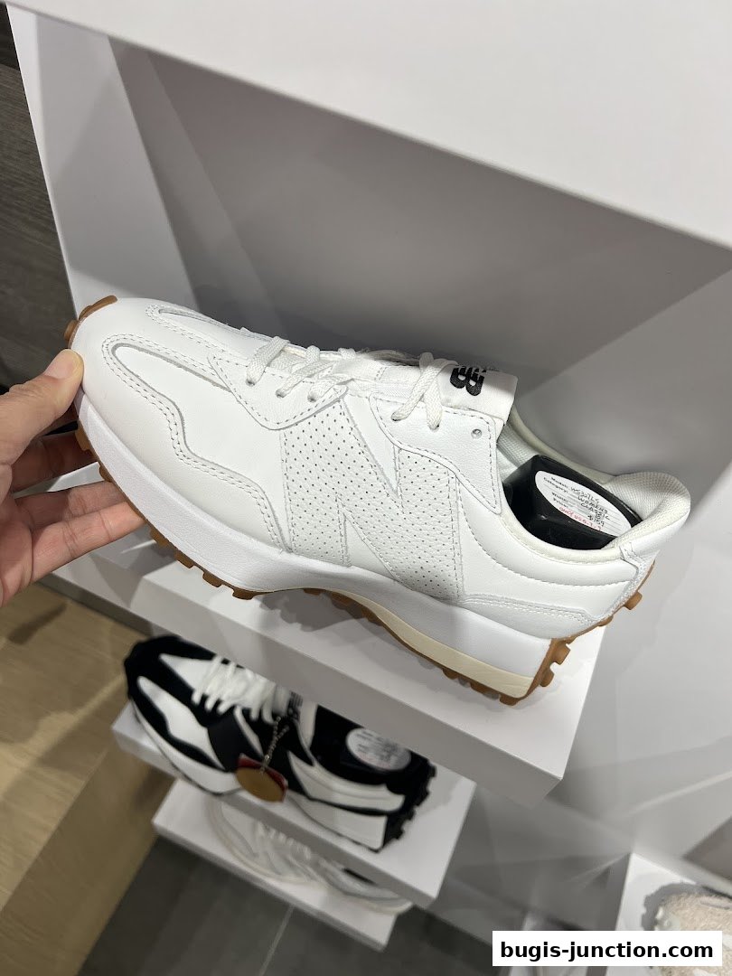 A hand holds a white New Balance sneaker with a gum sole, displayed on a shelf.