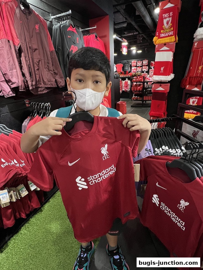 A young fan proudly holds a Liverpool FC jersey inside a brightly lit retail store.