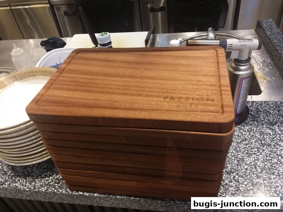 A stack of wooden cutting boards with the PAZZION cafe logo, next to plates and kitchen equipment.