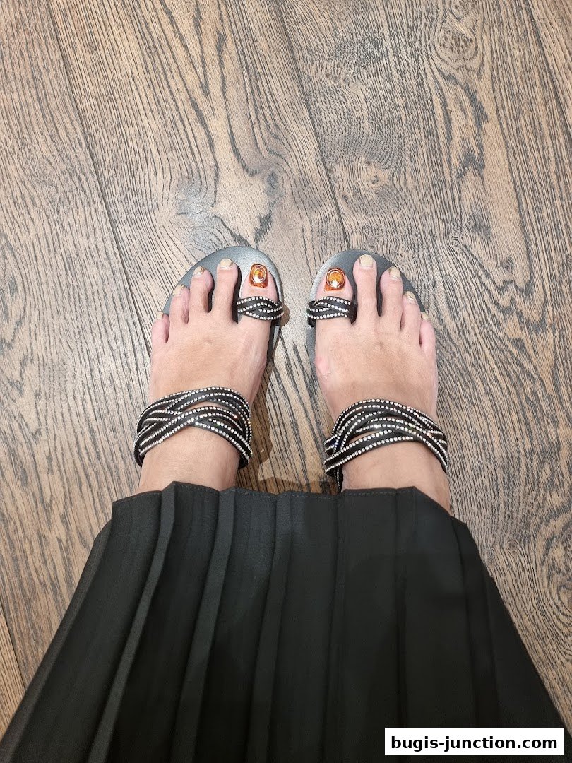 A pair of feet wearing stylish sandals with sparkling embellishments and a black pleated skirt.