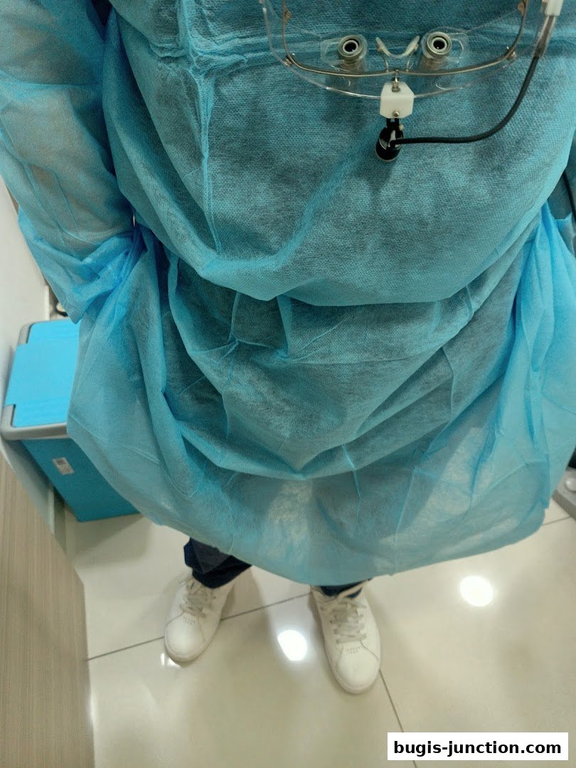 A person in a blue dental gown and loupes, standing in a clinic.