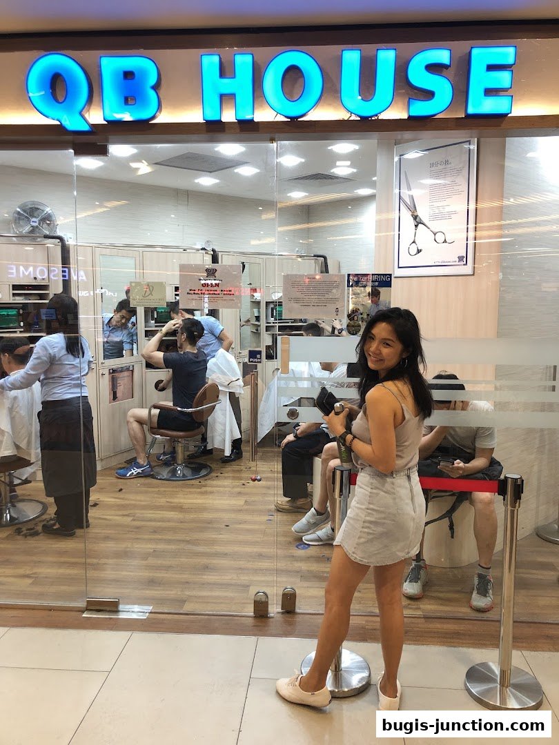 A woman poses in front of QB House salon, showing the interior with customers getting haircuts.