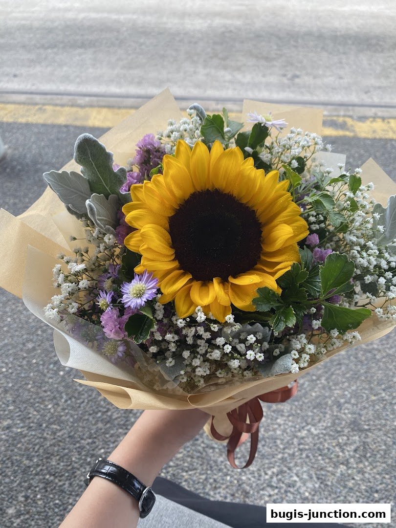 A hand holds a beautiful bouquet featuring a large sunflower and delicate filler flowers.