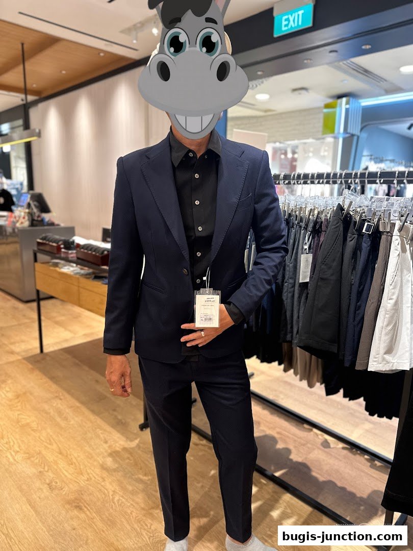 A person in a navy suit stands in a clothing store, with racks of pants visible.