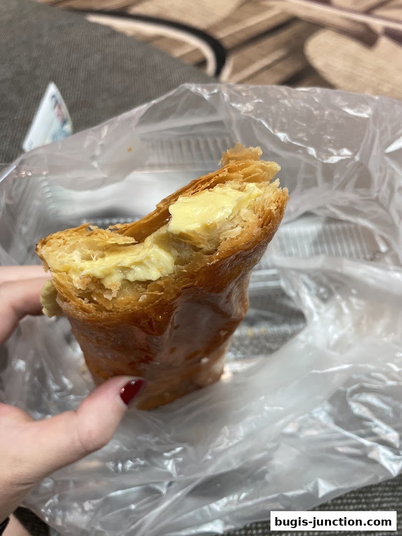 A hand holds a flaky pastry filled with creamy custard, packaged in plastic.