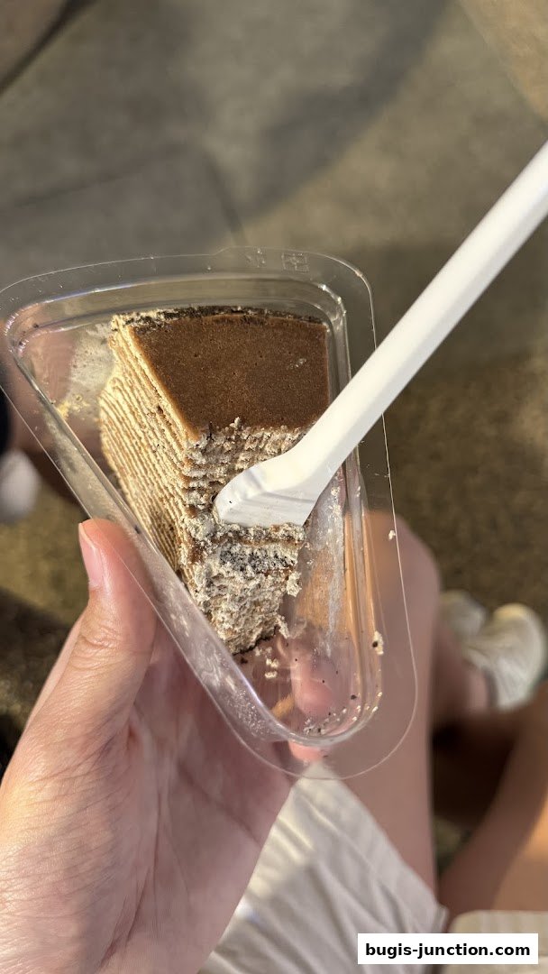 A hand holds a slice of layered chocolate cake with a fork in a clear plastic container.
