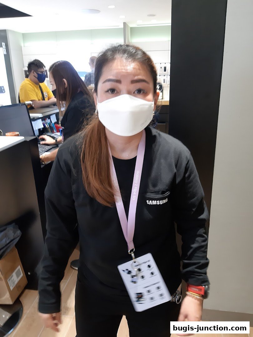 A Samsung store employee wearing a mask and a Samsung branded shirt stands in the store.