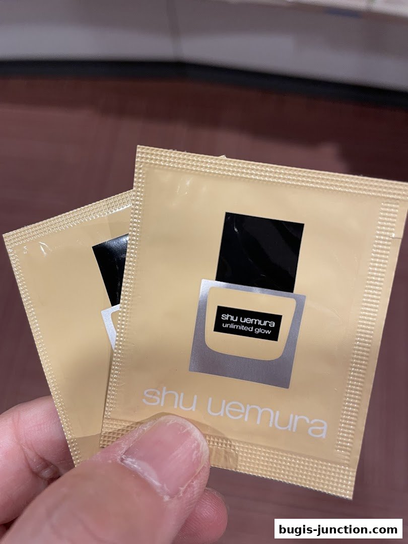 A hand holds two sample sachets of Shu Uemura Unlimited Glow foundation.