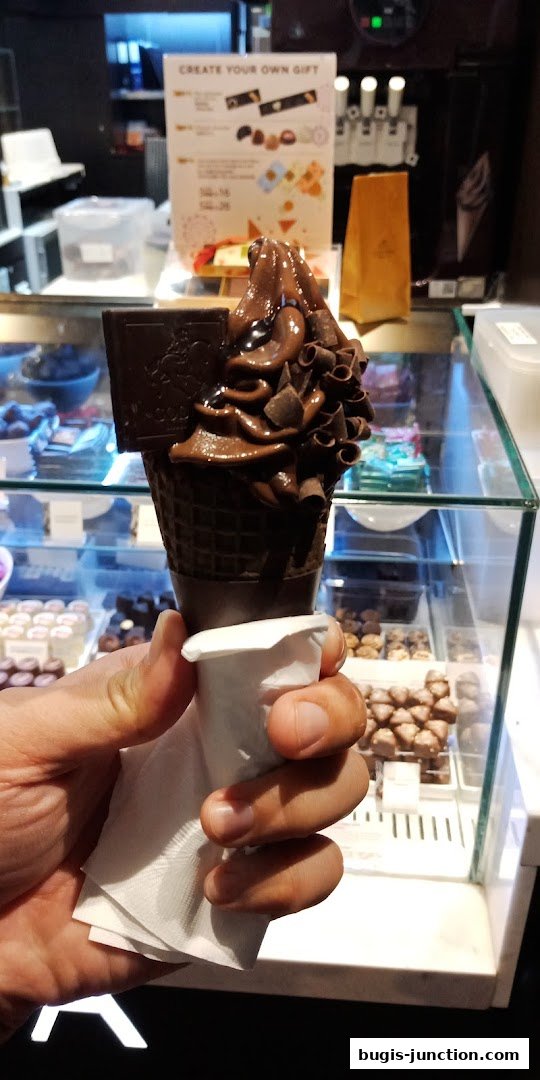 A hand holds a decadent chocolate soft serve ice cream cone with chocolate shavings.