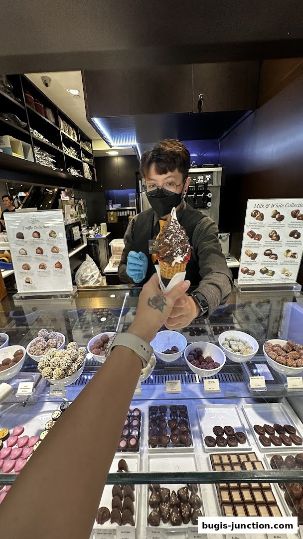 A person receives a chocolate ice cream cone from a Godiva shop employee.