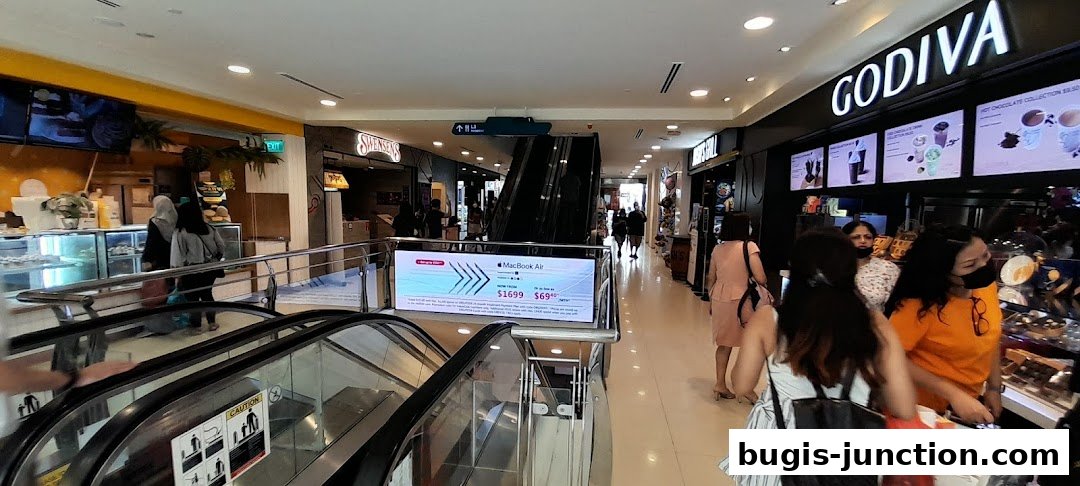 People shopping at Godiva and other stores in a mall with escalators.