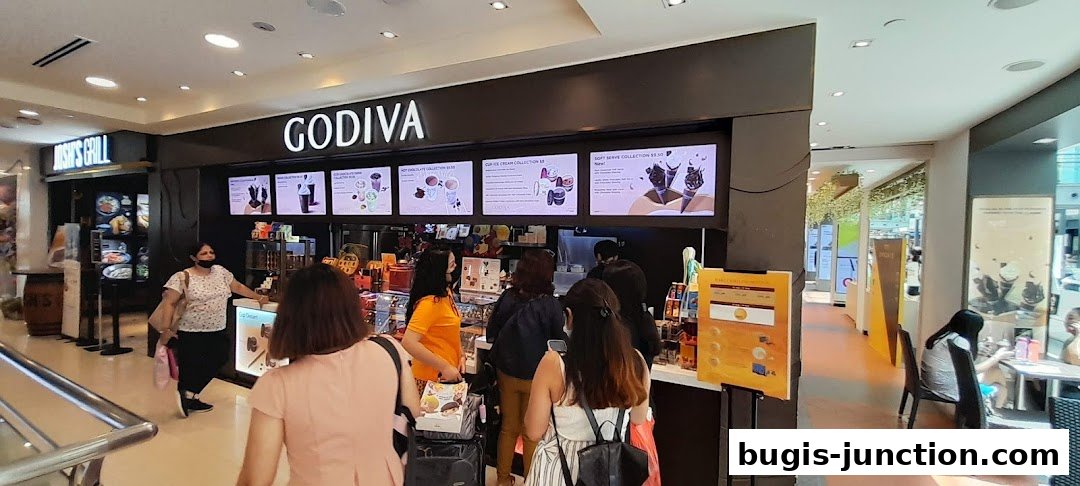 Customers queue at the Godiva chocolate shop, with illuminated menus displaying various chocolate and ice cream collections.