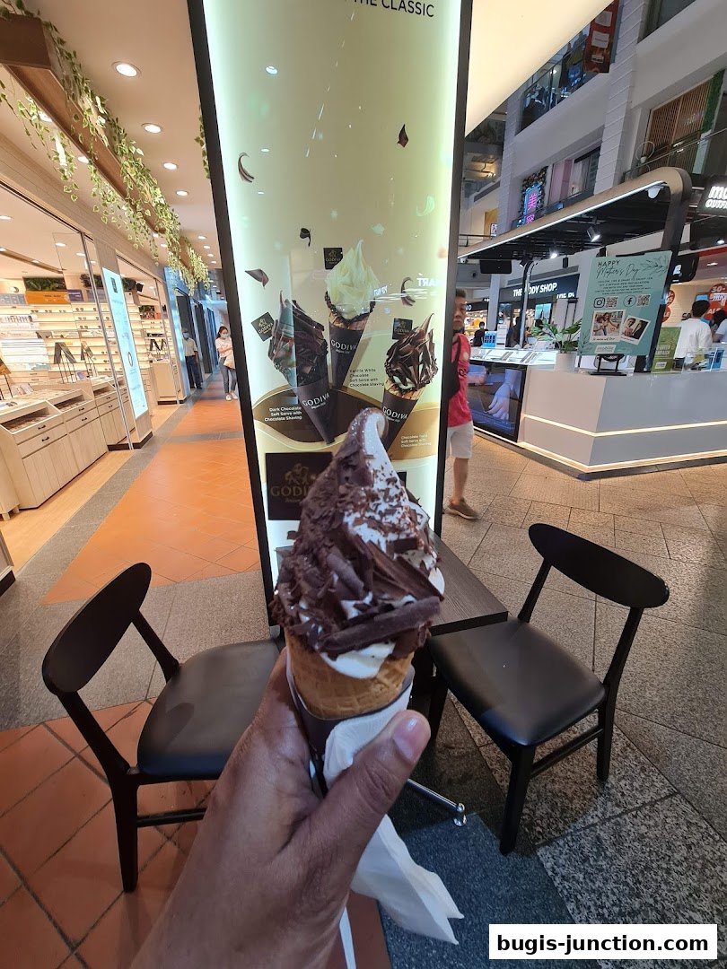 A hand holds a chocolate-covered ice cream cone in front of a Godiva display.