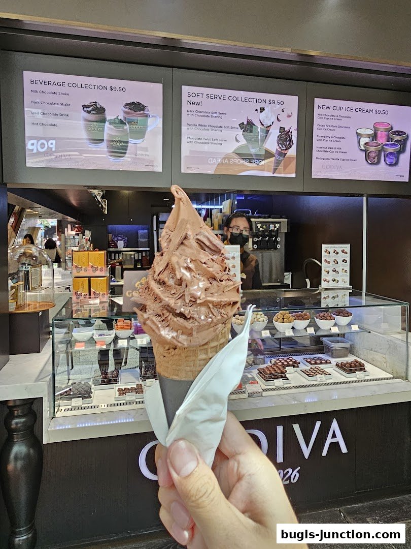 A hand holds a large chocolate soft serve cone in front of a Godiva shop display.