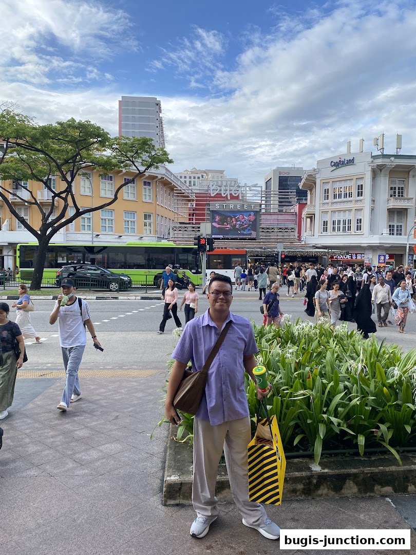 A man stands on a sidewalk in front of a busy street with shops and a bus.