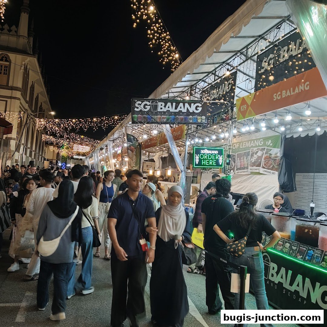 A bustling night market scene with many people walking past food stalls decorated with lights.