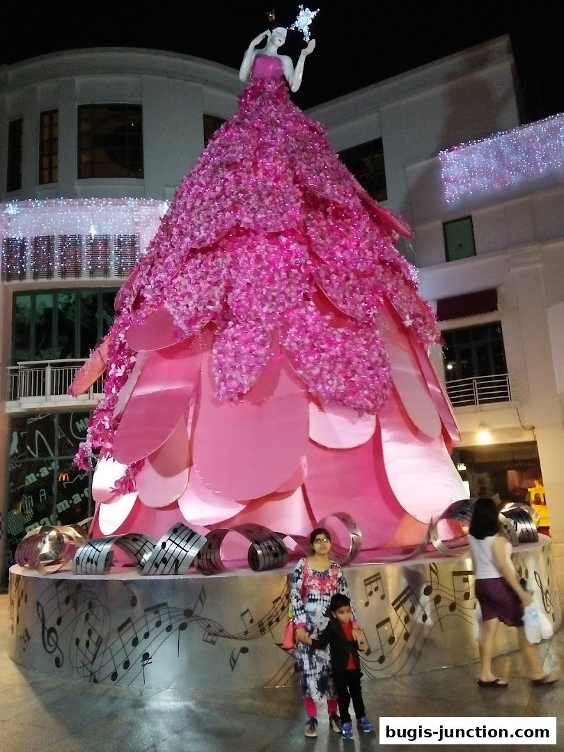 A festive pink Christmas tree with a doll on top, decorated with lights and musical notes.
