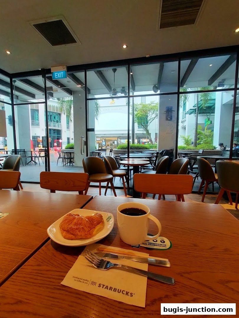 A table setting at Starbucks with a croissant, coffee, and cutlery, overlooking the mall.
