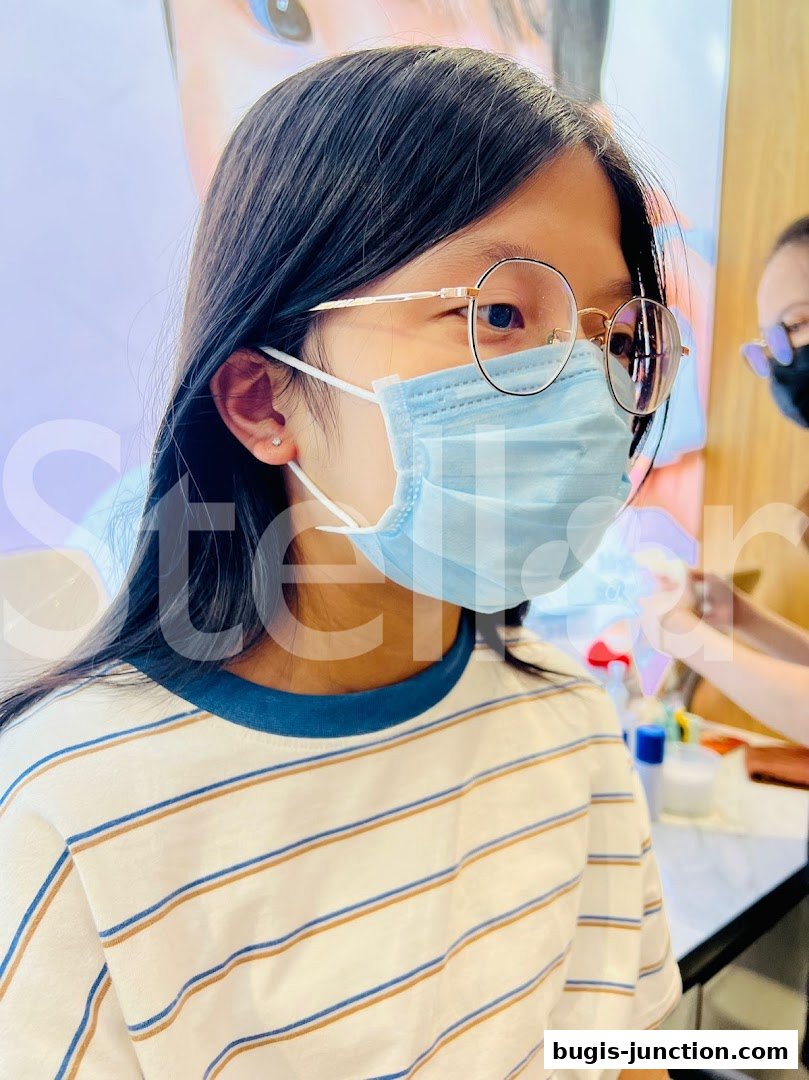 A young girl with glasses and a mask gets her ear pierced with a small stud.