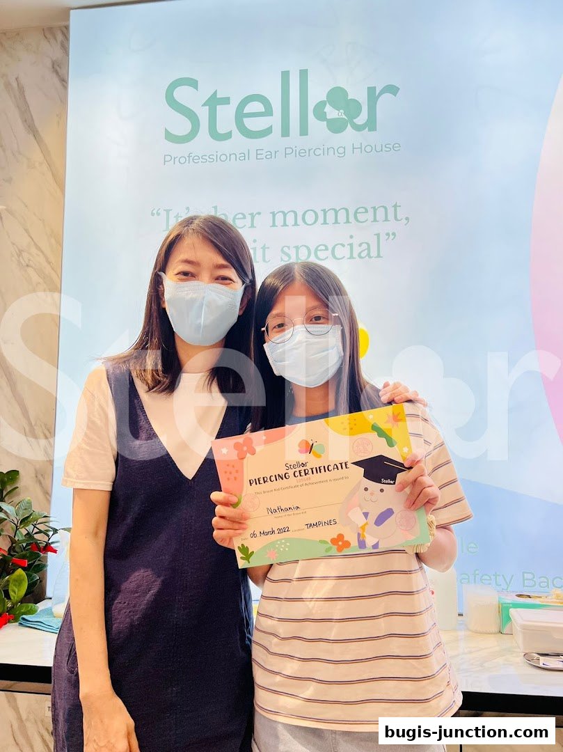 Two people, one holding a piercing certificate, pose in front of a Stellar Professional Ear Piercing House backdrop.