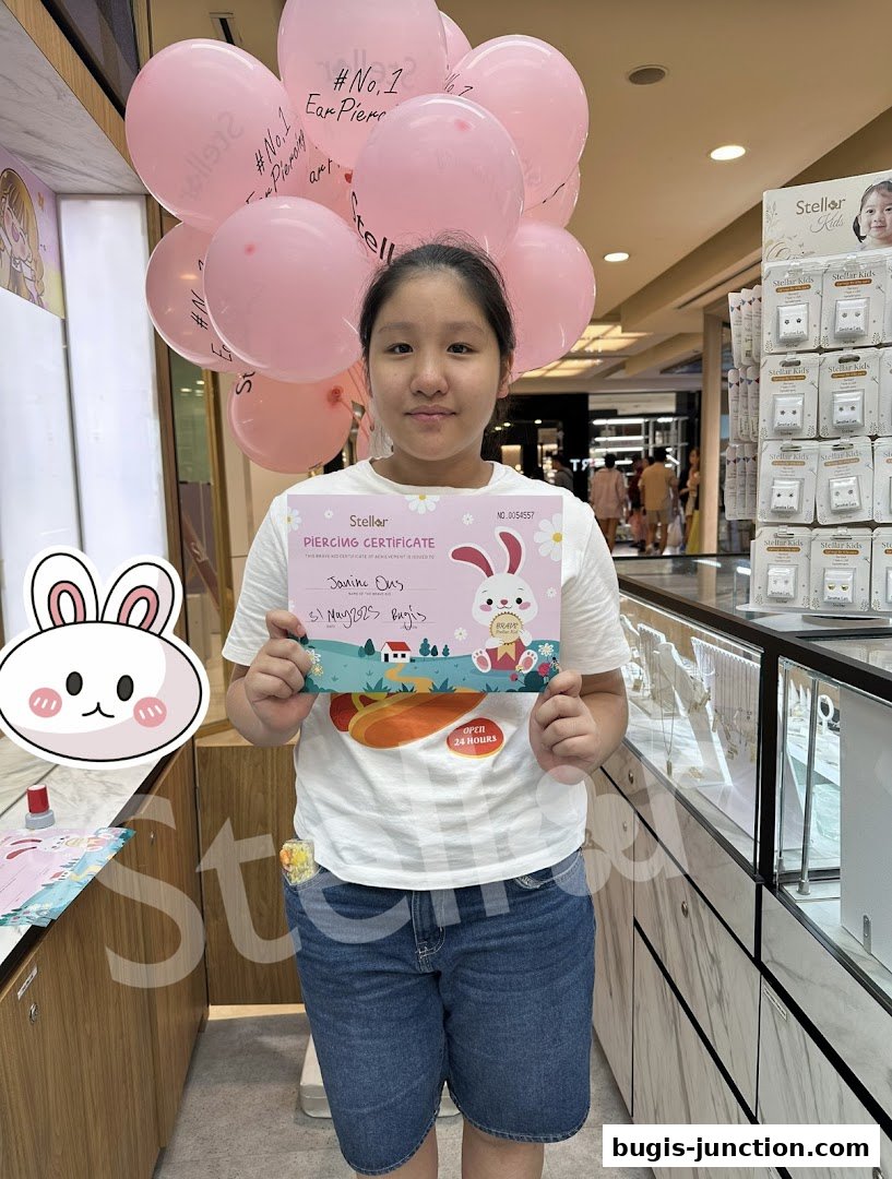 A young girl holds a piercing certificate with pink balloons behind her at Stellar.