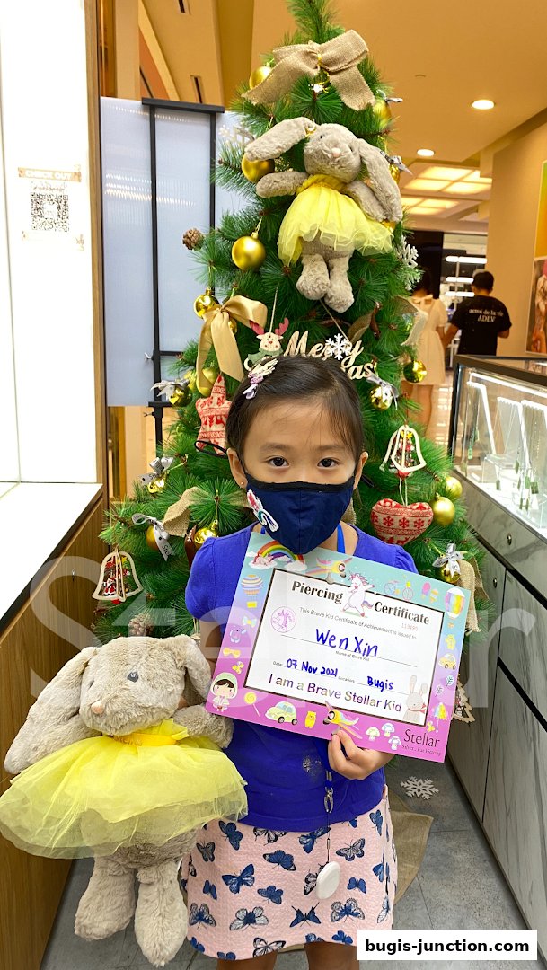 A young girl holds a piercing certificate and a stuffed bunny in front of a decorated Christmas tree.