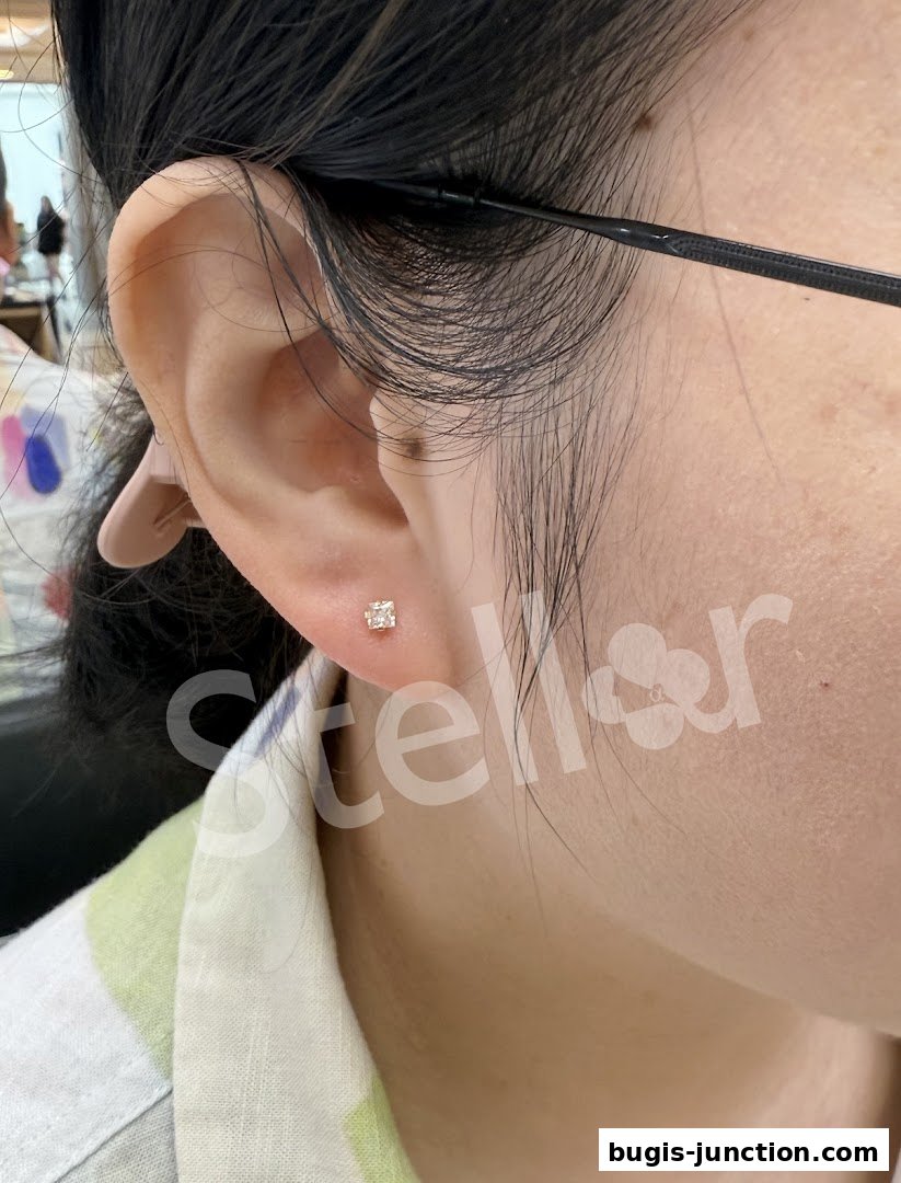 Close-up of a person's ear with a small, square stud earring, featuring a watermark.