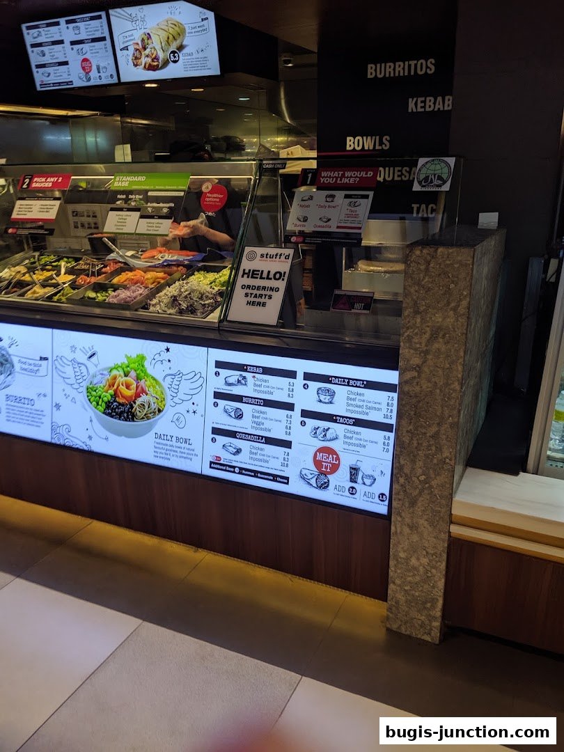 A food counter with a display of fresh ingredients and a digital menu board showing various meal options.