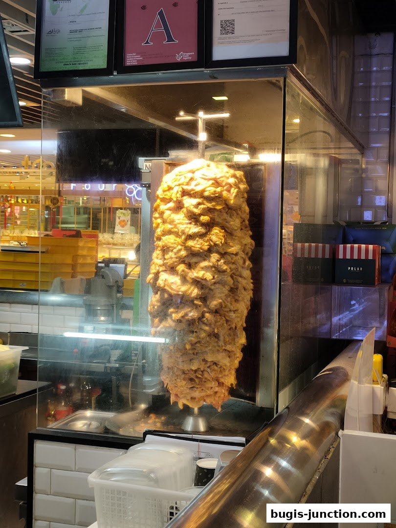A vertical rotisserie of seasoned meat is displayed in a glass enclosure at a food stall.