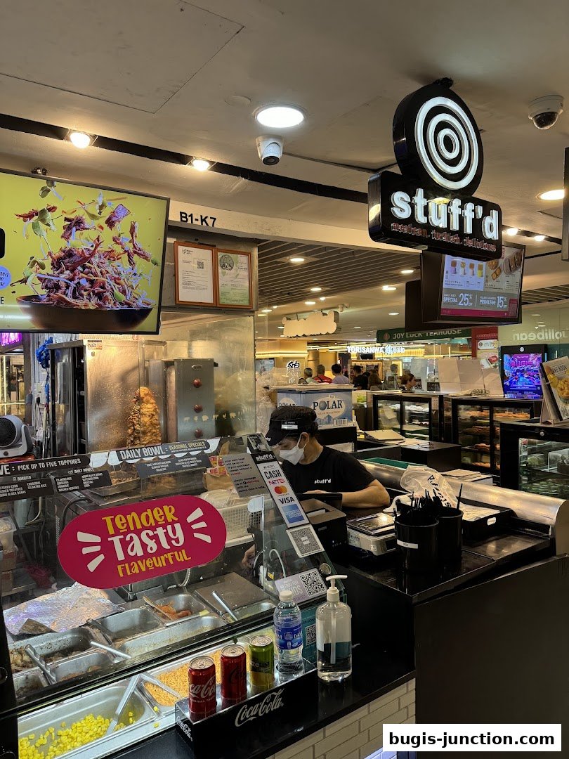 A food stall with a worker preparing food behind a counter, featuring a display of ingredients and drinks.