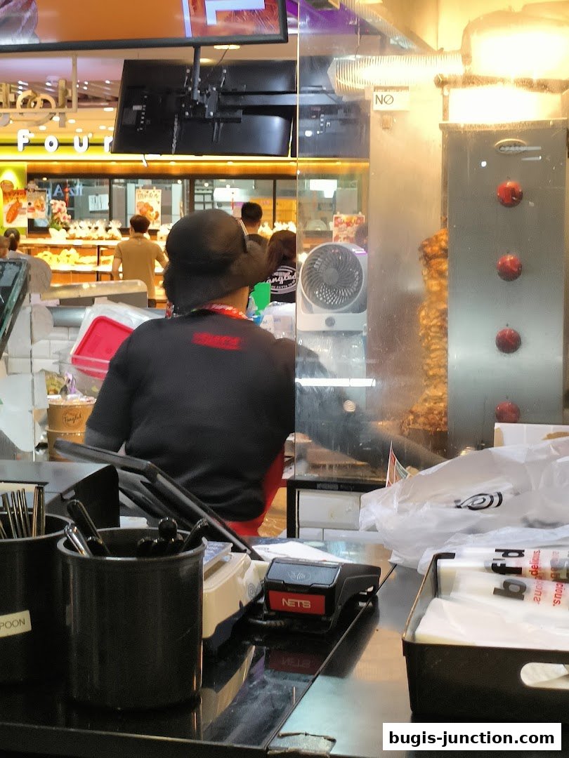 A view of a food stall counter with staff preparing food and a vertical rotisserie.