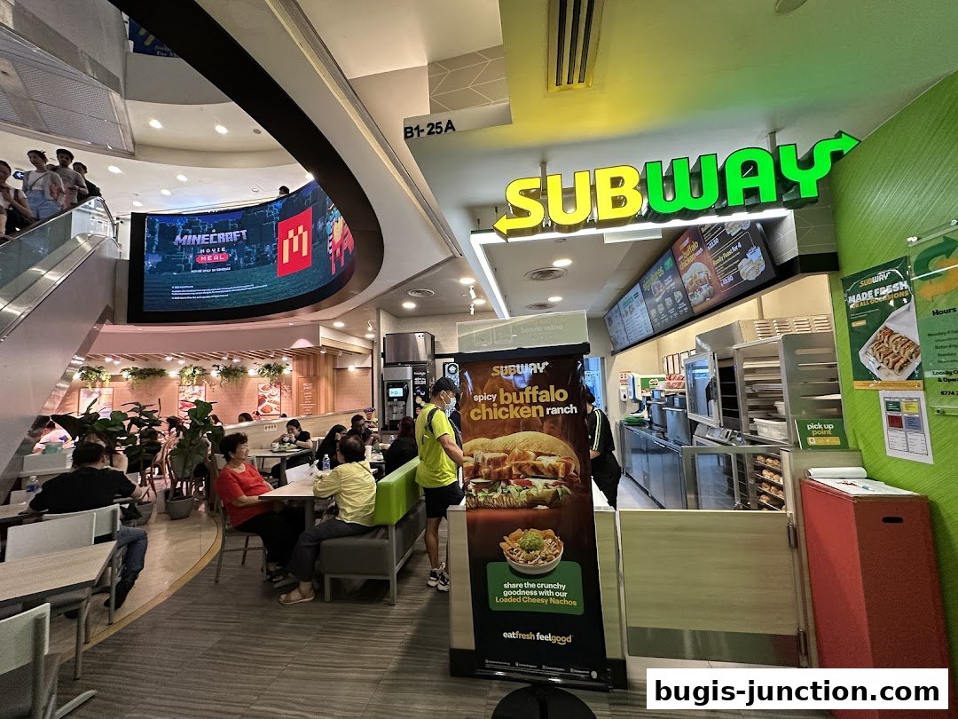 A Subway restaurant interior with customers and a large digital screen displaying a Minecraft movie advertisement.