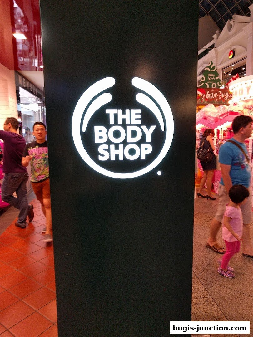 The Body Shop logo displayed prominently on a dark panel with shoppers in the background.