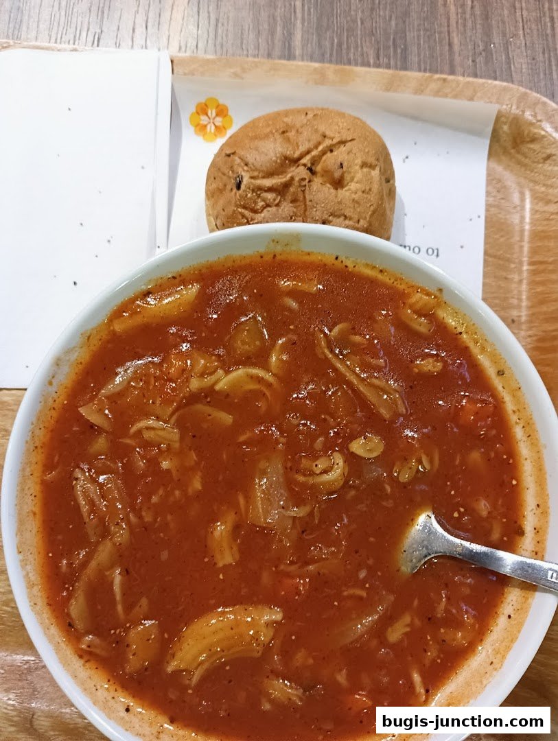 A bowl of hearty tomato pasta soup with a side of bread roll on a wooden tray.